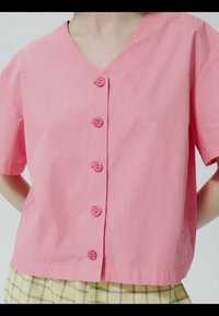 Personne portant un haut rose à manches courtes avec des boutons, associé à un pantalon à carreaux jaune, debout les mains derrière le dos.