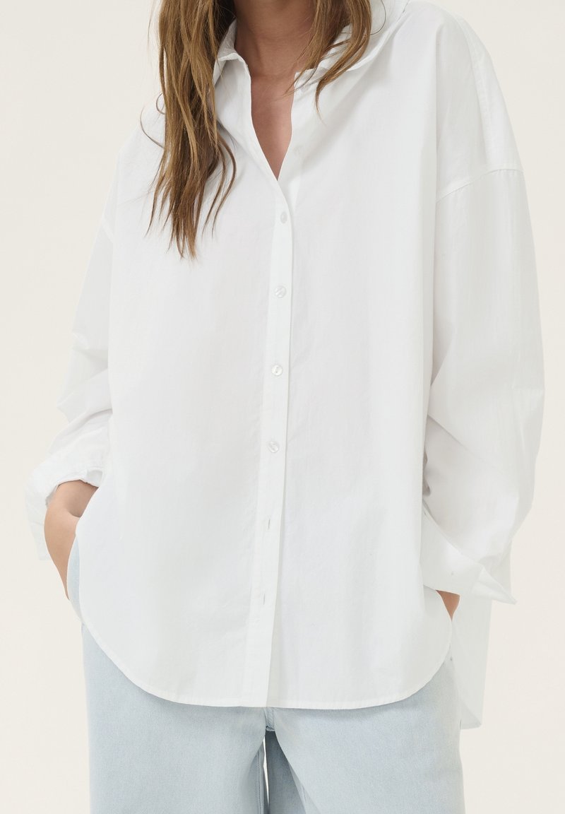 Woman wearing loose white button-up shirt with collar and light blue jeans, hands in pockets, against a plain light background.