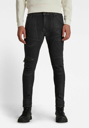 Mężczyzna noszący czarne, dopasowane, postrzępione jeansy z łatami na kolanach i suwakiem na lewym kolanie, czarne buty i beżową bluzę.