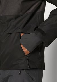 Main gantée noire glissant dans la poche zippée d'une veste noire et gris foncé avec une bande Velcro sur la manche.