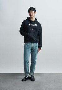 Sweat-shirt à capuche noir avec "Moschino Milano" imprimé en blanc, associé à un jean droit bleu clair et des chaussures noires.