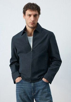 Man with hands in pockets wearing a dark zip-up jacket over a white shirt and blue jeans, standing against a plain light background.