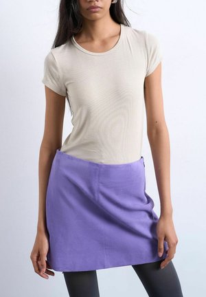 Woman wearing a beige ribbed short-sleeve top and a high-waisted purple skirt, standing against a plain light background.