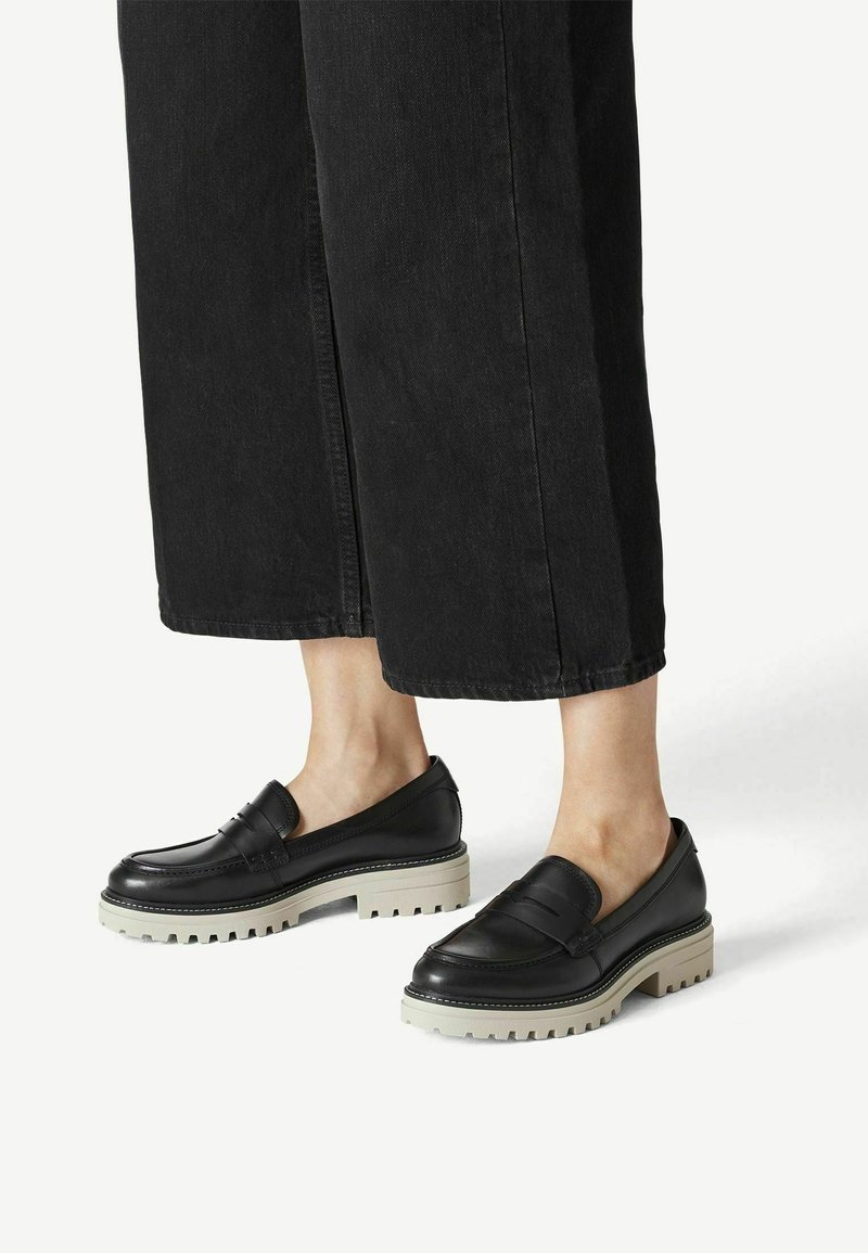 Pieds portant des mocassins en cuir noir avec des semelles blanches épaisses, associés à un pantalon noir à jambes larges et coupé, sur un fond blanc.