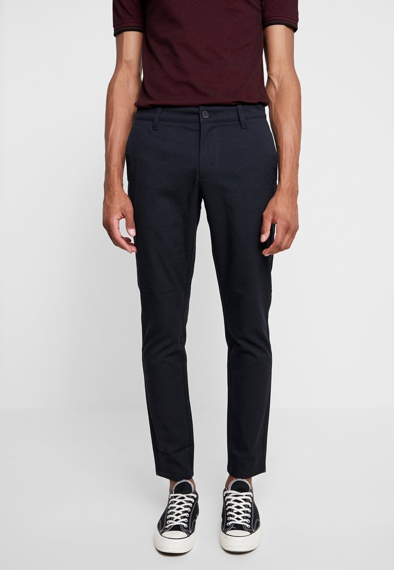 Pantalon bleu marine à coupe slim, doté d'une fermeture par bouton et de passants pour ceinture. Porté avec des baskets noires et blanches, sur un fond uni.