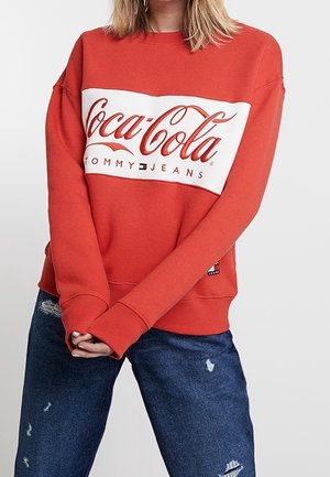 Nainen, jolla on päällään punainen Coca-Cola x Tommy Jeans -collegepaita ja kulutetut siniset farkut, seisoo kädet edessä ristissä.