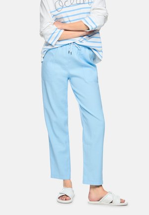 Person wearing light blue drawstring pants, white and blue striped long-sleeve shirt, and white braided slide sandals, standing with arms crossed.