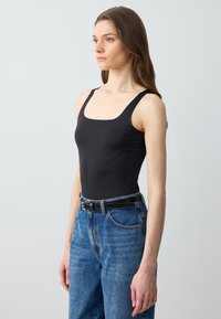 Bodysuit nero senza maniche con scollatura quadrata, abbinato a jeans blu a vita alta e una cintura nera, su uno sfondo neutro.