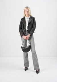 Chaqueta de motorista de cuero negro, camisa blanca de botones, jeans acampanados grises y bolso negro con studs, con tacones negros de punta, de pie contra un fondo blanco.