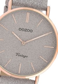 Montre avec un cadran argenté texturé et des accents en or rose. Présente des marqueurs d'heure minimalistes, le nom de la marque "OOZOO", et le mot "Vintage". Bracelet ajustable.