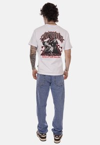 Uomo con t-shirt bianca con grafica di cane ringhiante, testo "PROPAGANDA" e "ENTRA A TUO RISCHIO", jeans blu larghi e sneakers Nike marroni.