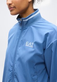 Κοντινό πλάνο ατόμου που φορούσε μπλε ζακέτα με φερμουάρ και με το λογότυπο EA7 Emporio Armani στο αριστερό στήθος και λεπτομέρειες ριγέ στο γιακά.