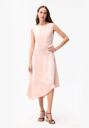 Kobieta z długimi brązowymi włosami, ubrana w asymetryczną, bezrękawną, bladoróżową sukienkę i brązowe sandały na wysokim obcasie, stojąca z jedną ręką na biodrze.