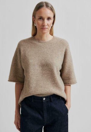 Hellbrauner Wollmix-Pullover mit Rundhalsausschnitt und kurzen Ärmeln, kombiniert mit dunklen Jeans mit vorderem Knopfverschluss.