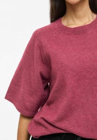 Sieviete, kas valkā šauru, apaļu kakla izgriezumu un mīkstu džempera džemperi dziļā rozā krāsā, kombinēta ar melnām biksēm uz vienkārša balta fona.