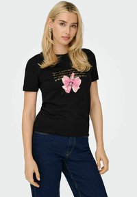 Kvinde med langt blondt hår, der bærer sølvfarvede hoops, en sort T-shirt med grafisk print af pink læbestift og sløjfe, samt mørkeblå jeans.