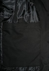 Wnętrze czarnej kurtki wykonane z gładkiego materiału, z dyskretną kieszenią i teksturowaną podszewką. Minimalistyczny design z naciskiem na funkcjonalność.