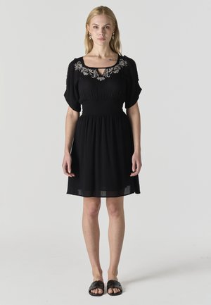 Mujer con un vestido negro hasta la rodilla con escote bordado de flores y sandalias negras sin cierre, de pie frente a un fondo liso.