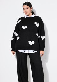 Schwarzer gestrickter Pullover mit weißen Herzmustern, übergroßer Schnitt, runder Hals, über einem weißen Hemd mit Kragen getragen. Strukturierter und weicher Stoff.