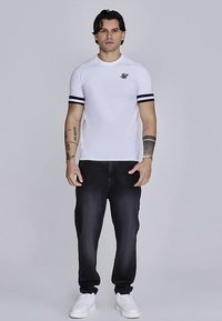 Bela majica s kratkimi rokavi s črnimi črtami na rokavih, kombinirana s temnimi kavbojkami in belimi supergami. Na prsih je majhna nalepka z logotipom.