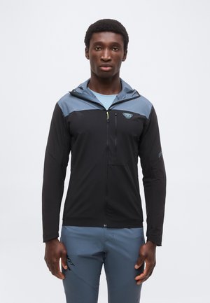 Νέος άνδρας που φοράει μαύρο-μπλε μπουφάν με φερμουάρ και λογότυπο στο στήθος, στέκεται μπροστά σε ένα απλό λευκό φόντο.