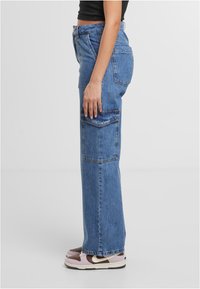 Pantalones cargo de mezclilla azul con un corte holgado, que presentan grandes bolsillos laterales y un diseño de pierna recta, combinados con zapatillas marrones y rosas.
