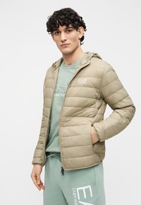 Jachetă matlasată verde măsliniu deschis cu glugă, având un design matlasat și buzunare laterale. Purtaă peste o cămașă verde mentă cu un logo.