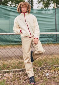 Beige fleece jacket with red accents and zipper, paired with beige pants. Black shoes feature pink details, set against a green background.
