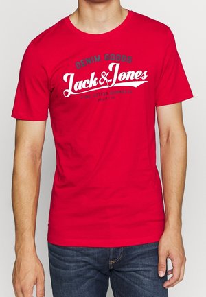 Man wearing red short-sleeve T-shirt with "Denim Goods Jack & Jones Fine Cotton Garments" text, paired with dark blue jeans.