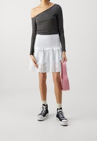 Grijze top met lange mouwen en open schouders, witte katoenen rok met meerdere lagen en geborduurde zoom, zwarte hoge sneakers, met een roze handtas.