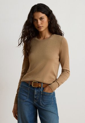 Femme aux longs cheveux bouclés foncés portant un pull camel ajusté, un jean bleu et une ceinture en cuir marron avec une boucle dorée, debout, une main dans la poche.