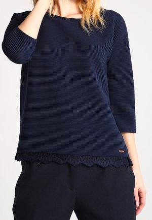 Vrouw die een marineblauwe geribde top draagt met driekwartmouwen en een kanten rand, gecombineerd met een zwarte broek.