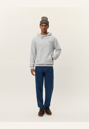 Gray hoodie with a front pocket, featuring a small logo. Paired with blue denim jeans and brown shoes, complemented by a brown beanie.