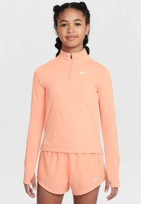 Długorękawowy top w jasnobrzoskwiniowym kolorze z białym logo Nike i suwakiem do ćwiartki, zestawiony z pasującymi jasnobrzoskwiniowymi szortami, miękki materiał, dopasowany fason.