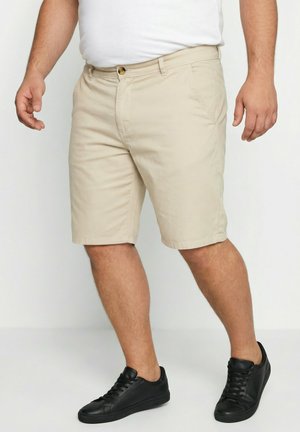 Mand iført beige knælange shorts, hvid skjorte og sorte lave sneakers, stående på en ensfarvet hvid baggrund.