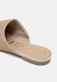 Beige mockasins i mocka med öppen tå och låg träklack; har en slät inredning och en dekorativ gyllene logotyp på sulan.