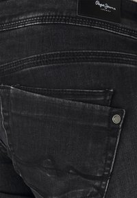 Jeans de mezclilla negra con un bolsillo trasero con un botón metálico, textura desgastada sutil y costuras en contraste. Etiqueta de Pepe Jeans en la cintura.