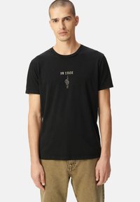 Camiseta de algodón negra con cuello redondo, que presenta un estampado gráfico de una mano y el texto "EN ESCENA" en blanco arriba. Mangas cortas y corte estándar.