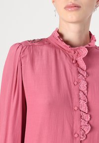 Pink blouse with a ruffled collar, decorative lace trim, and button closure, featuring textured fabric and bead accents on the shoulders.