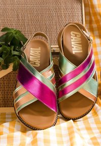 Sandales marron avec des brides métalliques croisées vertes, roses et pêche, étiquetées « CAVAL », posées sur un tapis tissé et un tissu à carreaux jaune.