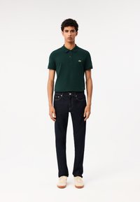 Polo de color verde oscuro con mangas cortas y cuello, combinado con jeans de color azul oscuro y zapatillas de tonos claros. Presenta el logo de Lacoste en la camisa.