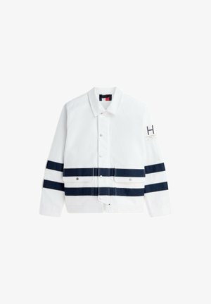 Veste blanche avec des rayures horizontales bleu marine sur les manches et à la taille, fermeture à pression devant, col et petit écusson logo sur le bras gauche.
