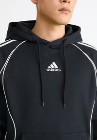 Homem a usar um hoodie preto da Adidas com o logotipo branco, cordões e detalhes em branco nos ombros, contra um fundo simples.