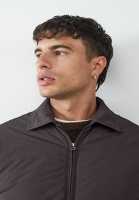 Dunkelgraue, leichte Zip-Jacke mit spitzem Kragen, getragen über einem braunen Pullover. Verfügt über einen silbernen Reißverschluss und eine glatte Textur.