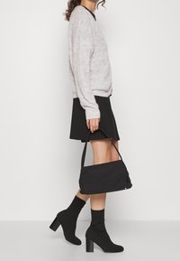 Zwarte schoudertas van stof, met een zachte textuur en een minimalistisch ontwerp. Gedragen bij een lichtgrijs gebreid sweater en een zwarte rok.