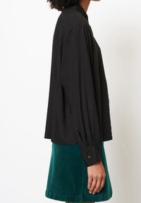 Schwarze Bluse mit langen Ärmeln, strukturiertem Kragen, gefältelten Ärmeln und geknöpften Manschetten, kombiniert mit einem petrolfarbenen Cordrock.