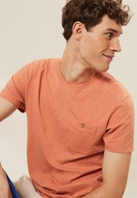 Kurzärmliges T-Shirt mit Rundhalsausschnitt in hellem Koralle mit strukturierter Oberfläche, ausgestattet mit einer Brusttasche auf der linken Seite und einem kleinen Knopfakzent.