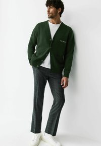 Groen gebreid vest met V-hals, knoopsluiting en twee voorzakken, gecombineerd met grijze pantalon met patroon en witte sneakers.