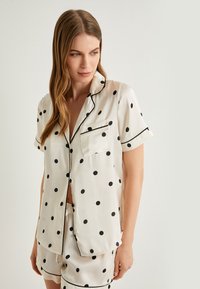 Valkoisesta silkistä valmistettu pyjama-setti, jossa on mustia kokardiakuviota, lyhyet hihat, rintatasku ja musta kanttaus kauluksessa sekä shortseissa.