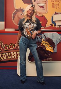 Camiseta gráfica que presenta a un personaje a caballo, jeans de mezclilla acampanados azules, bolso de cuero marrón con una bufanda amarilla de patrón.